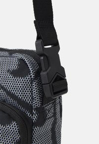 Černobílá vzorovaná taška přes rameno z odolné tkaniny, s nastavitelným popruhem a bezpečnou plastovou sponou na boku.