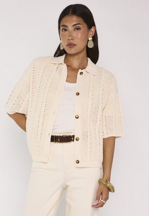 Donna che indossa un cardigan corto in maglia crema con bottoni, top bianco, pantaloni crema, cintura marrone, grandi orecchini e braccialetti a un polso.