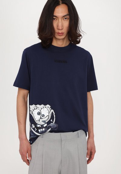Mørkeblå bomulds T-shirt med et trykt grafisk motiv af en tegneseriefigur på venstre side; har en ribbet rund hals og en afslappet pasform.