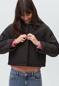 Jachetă scurtă, matlasată, de culoare neagră, cu deschidere frontală cu nasturi, având guler și țesătură texturată, purtată peste un pulover roz, asociată cu blugi albaștri.