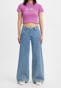 Person klädd i en rosa kort t-shirt med texturerad yta och färgglad text, höga vida blå jeans och sneakers med grön-vita rutor.