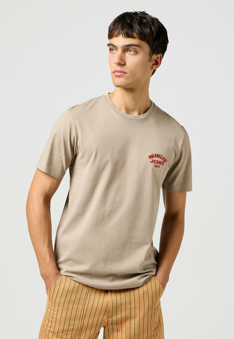Beige bomull t-shirt med kort ärm och rundad halslinning, med röd broderad logotyp "Wrangler Jeans 1947" på bröstet.