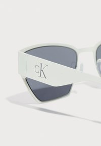Hvite solbriller med geometrisk form, grå linser og en fremtredende cK-logo på siden, med en matt finish og slank design.