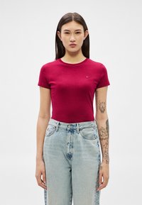 Jauna sieviete ar gariem tumšiem matiem, tērpusies sarkanā cieši pieguļošā T-kreklā un gaiši zilās augsta vidukļa džinsās, rāda tetovējumus uz kreisās rokas.