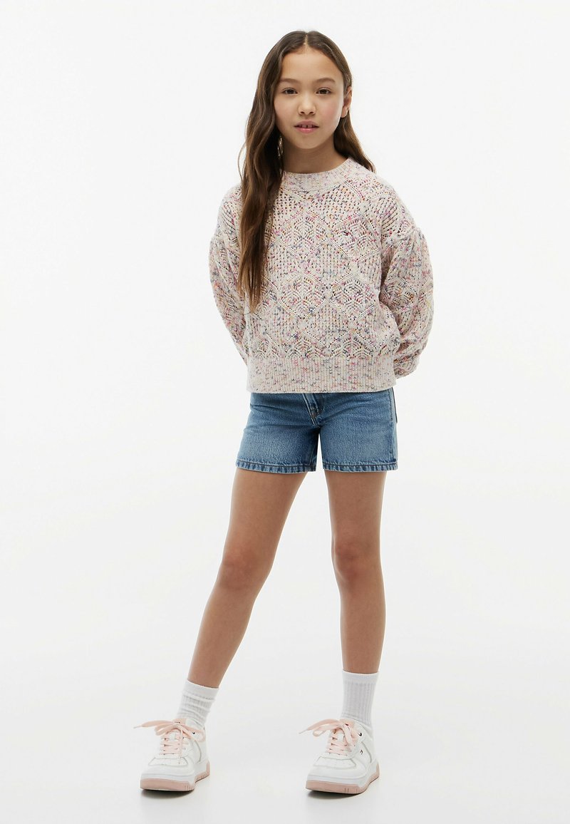 Ung pige stående med hænderne bag ryggen iført en mønstret striktrøje, denimshorts, hvide sokker og hvide sneakers.