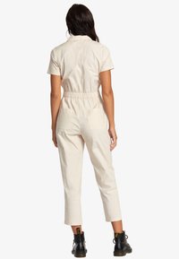 Beige jumpsuit with short sleeves, elastic waist, and two back pockets. The fabric appears lightweight and smooth, paired with black boots.