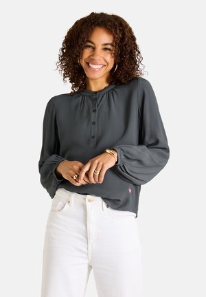 Lächelnde Frau mit lockigem Haar, die eine dunkelgraue geknöpfte Bluse und weiße Hose trägt, ergänzt durch goldene Ringe und Armbänder.