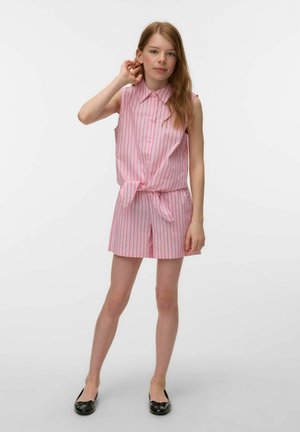 Teenagepige står og har en pink og hvid stribet ærmeløs skjorte med krave bundet ved taljen, matchende shorts og sorte balletsko.