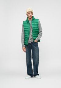 Green padded vest over a gray long-sleeve shirt, paired with blue wide-leg jeans and white sneakers. The model wears a beige knit beanie.