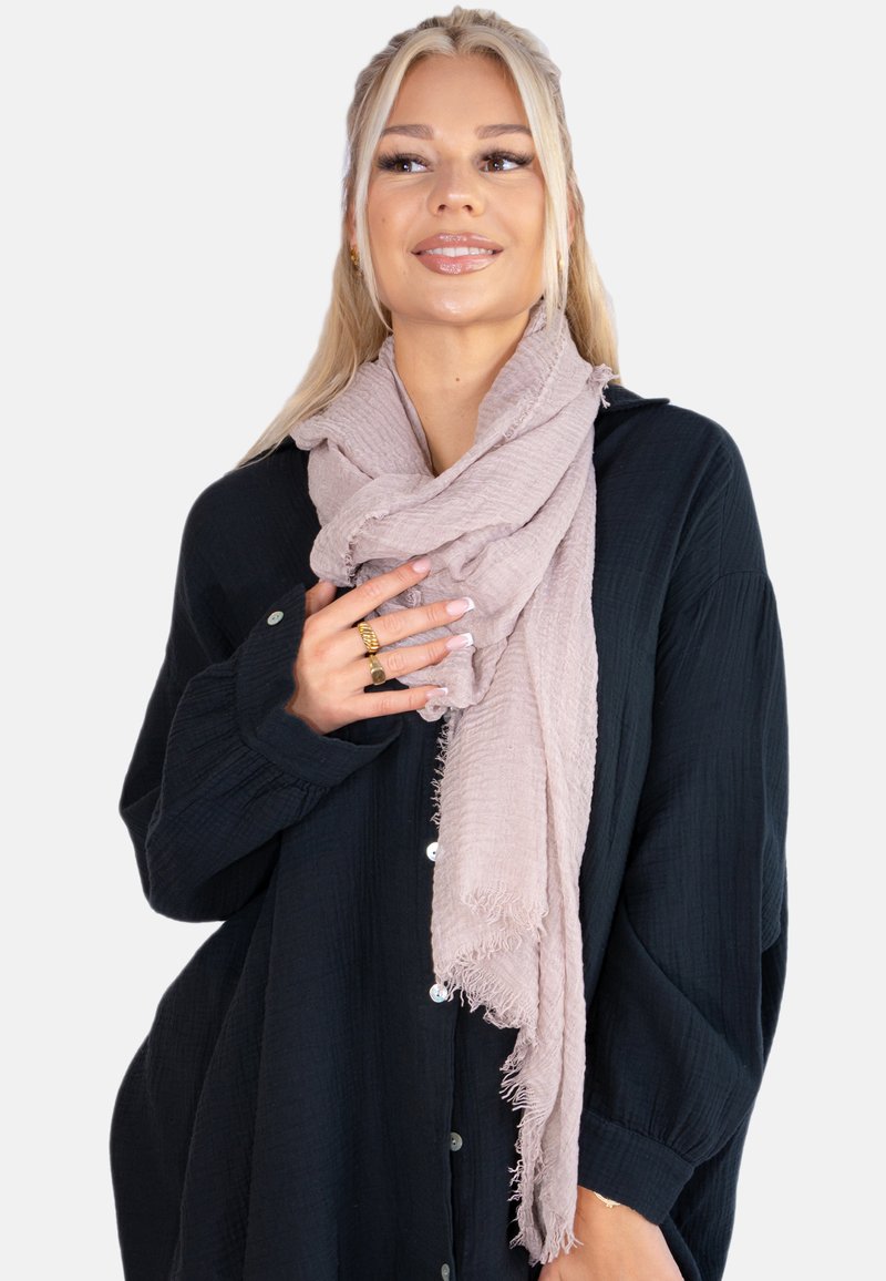 Écharpe rose clair, texturée, tombant librement autour du cou. Fabriquée en tissu doux avec des bords frangés, portée par-dessus une chemise à boutons sombre.