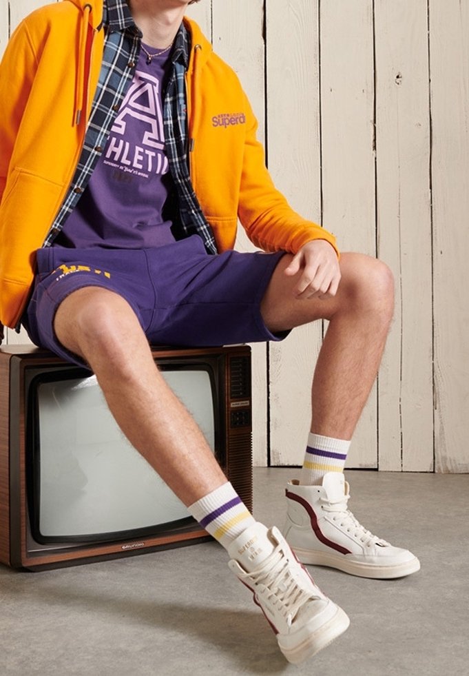 Personne portant un sweat à capuche orange, un short de sport violet et des baskets montantes blanches, assise sur une télévision vintage contre un mur en bois.