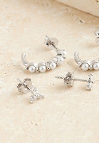 Silberne Creolen mit Perlakzenten und kleine Ohrstecker mit floralem Design und Edelsteinverzierungen auf einer strukturierten Oberfläche.