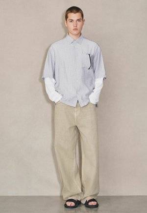 Junge Person trägt ein geschichtetes weißes Langarmhemd und ein kariertes Kurzarmhemd, beige weite Hosen, schwarze Sandalen, steht vor einer schlichten Wand.