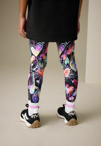 Farebné legíny s motívmi srdiečok v odtieňoch modrej, ružovej a žltej na čiernom pozadí, kombinované s čiernymi topánkami a pruhovanými ponožkami.