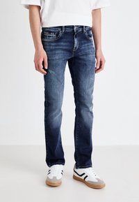 Men's dark blue jeans with light distressing, five-pocket design, subtle fading, and paired with white sneakers featuring black accents.