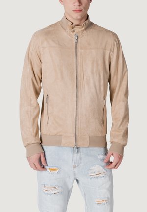 Veste en suède beige avec col montant, fermeture éclair frontale et poignets côtelés. Les détails notables incluent des poches latérales zippées et une texture lisse.
