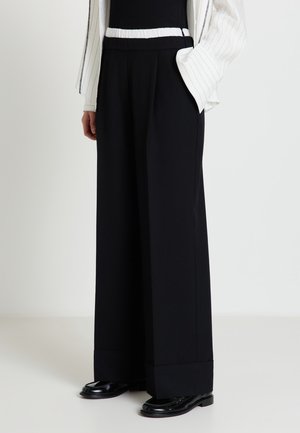 Persona che indossa pantaloni neri a gamba larga con elastico bianco in vita, scarpe nere e giacca bianca a righe sottili con la mano in tasca.