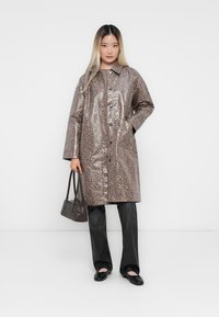 Leopard print, shiny vinyl trench coat, buttoned front, side pockets, layered over dark pants and flat shoes. Model holding a small handbag.