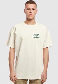 Camiseta de manga corta color crema hecha de algodón, con el texto verde "GAIN FAST LIVE WELL" y una pequeña impresión gráfica en el lado izquierdo del pecho.