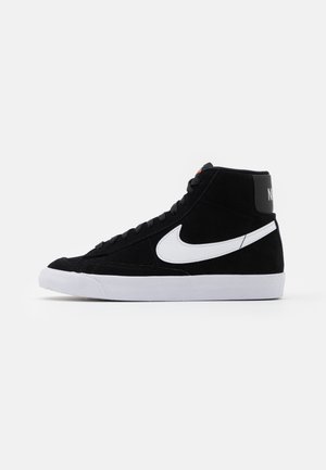 Czarny sneaker za kostkę z białym logo Nike po boku i białą podeszwą, pokazany w widoku z profilu na jasnym, jednolitym tle.