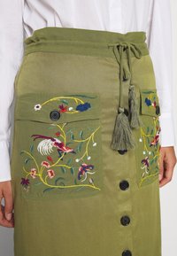 Falda verde olivo con patrones florales bordados, dos bolsillos frontales, cintura con cordón y botones negros en la parte delantera. Textura suave.