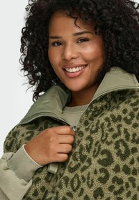 Smilende person med krøllete hår iført en olivengrønn fleecejakke med leopardmønster, delvis glidelås, med matchende indre krage.