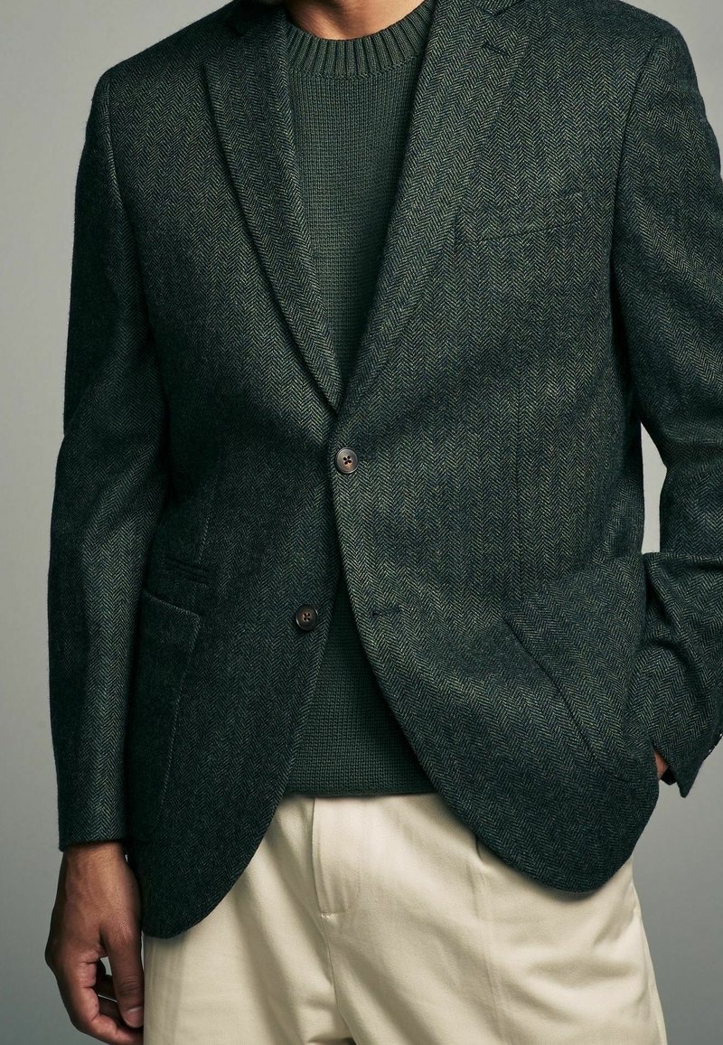 Homme portant un blazer vert foncé à chevrons sur un pull en tricot vert foncé et un pantalon beige clair, une main dans la poche.