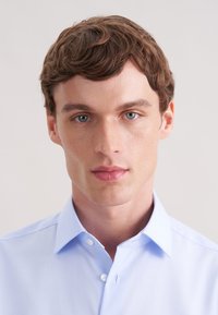 Jasnoniebieska koszula zapinana na guziki z klasycznym kołnierzykiem, wykonana z gładkiej tkaniny o subtelnym połysku. Model ma krótkie, falowane brązowe włosy i jasną karnację.