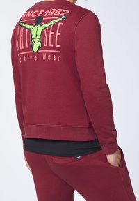 Sudadera marrón burdeos de mangas largas, con puños y dobladillo acanalados. Presenta un estampado gráfico en la parte posterior y un cuello redondo. Está hecha de una tela suave.