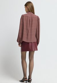 Langarmige, rood gebloemde blouse met volumineuze mouwen, gecombineerd met een bordeaux-kleurige neplederen minirok en zwarte haksandalen.