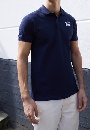 Mann trägt ein marineblaues Lacoste-Poloshirt und weiße Hosen, steht vor einem zweifarbigen Hintergrund aus Weiß und Dunkelgrau.