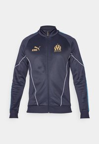 Chaqueta de chándal navy hecha de poliéster, con ribetes blancos, acentos del logo dorado y rayas azules en las mangas. Cierre frontal de cremallera completa.