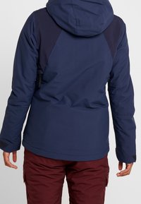 Giacca isolante blu navy con cappuccio, pannelli spalla scuri a contrasto e maniche testurizzate, abbinata a pantaloni bordeaux.