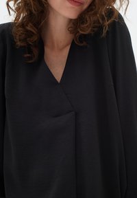Top nero largo con scollatura a V, tessuto morbido e un sottile dettaglio a piega diagonale sul fronte. Presenta maniche lunghe.