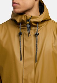 Giacca impermeabile color senape, con colletto alto, cordini regolabili neri e grigi e chiusura frontale con zip e tirazip con marchio.