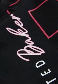 Musta kangaspaita, jossa on näkyvä vaaleanpunainen kirjailtu teksti "Baker" ja valkoiset painetut kirjaimet. Sileä pinta, jossa on kontrastoivia design-elementtejä.