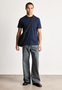 Homem vestindo uma t-shirt azul-marinho, jeans cinzentos largos, sapatos pretos e uma bolsa cruzada preta, de pé contra um fundo branco liso.