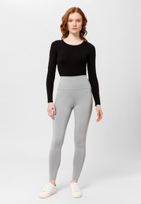Maglietta nera a maniche lunghe abbinata a leggings grigi chiari a vita alta. Sneakers bianche completano l'outfit. Sfondo semplice con toni neutri.