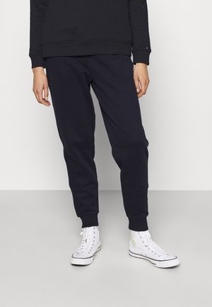 Personne portant un pantalon de jogging noir, un sweat-shirt noir et des baskets montantes blanches debout sur un sol gris contre un mur blanc.