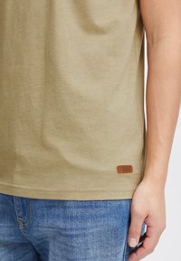 Khaki krāsa kokvilnas T-krekls ar īsām piedurknēm, ar rakstainu brūnu ādas birku ar uzrakstu "Solid" apakšējā kreisajā stūrī. Valkāts kopā ar ziliem džinsiem.