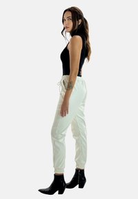 Schwarzes ärmelloses Oberteil, glänzende weiße Jogginghosen mit elastischen Bündchen, kombiniert mit schwarzen Stiefeletten. Seitenansicht vor neutralem Hintergrund.