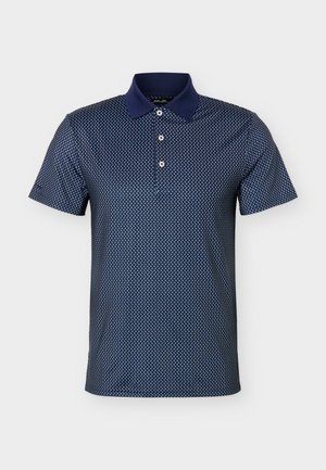 Mladý muž stojaci s rukami v vreckách, oblečený v námornícky modrý vzorovaný polokošeli, svetlošedých nohaviciach, bielej golfovej rukavici, čiernom opasku a bielych teniskách.