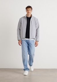 Lys grå, quiltet jakke, svart genser med rund hals, lyseblå jeans og hvite joggesko. Jakken har front uten glidelås og en avslappet passform.