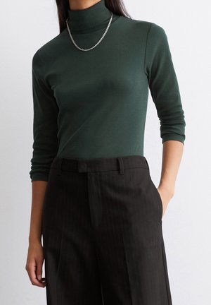 Personne portant un col roulé vert foncé à manches trois-quarts, un pantalon noir à fines rayures et un collier en chaîne argentée.