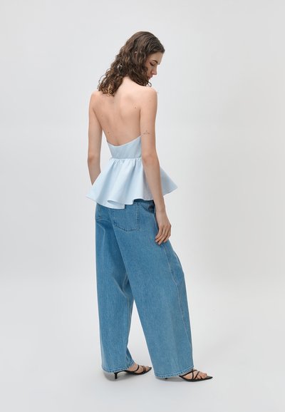 Femme aux cheveux bouclés portant un haut bleu clair sans dos avec basque, un jean bleu à jambes larges et des talons noirs à lanières, debout devant un fond uni.