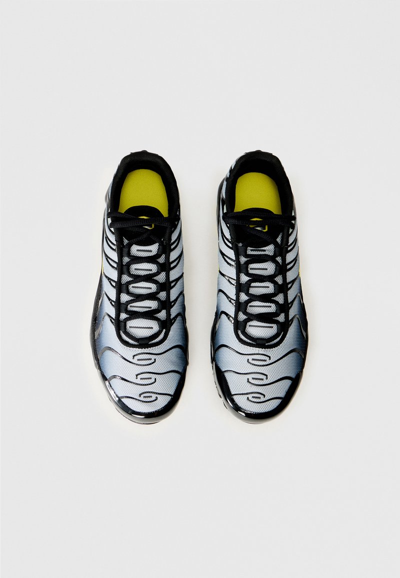 Ein Paar graue und schwarze Sportschuhe mit schwarzen Schnürsenkeln und gelben Einlegesohlen, von oben auf einem weißen Hintergrund betrachtet.