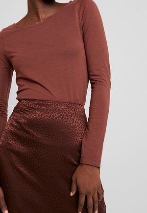 Vrouw draagt een lange mouw rusty-rode top en een glanzende donkere bruine rok met een subtiel luipaardprintpatroon.