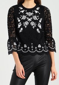 Svart spetsblus med vit blommig brodyr, vågig nederkant och ärmar, bärs med svarta läderbyxor av en stående person.