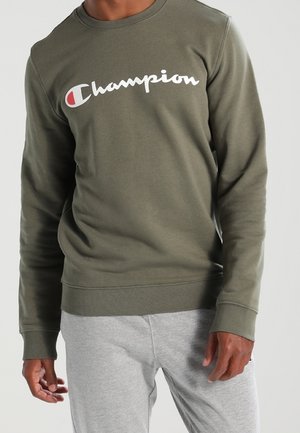 Mand iført en olivengrøn Champion sweatshirt med logo på tværs af brystet og lysegrå joggingbukser stående mod en ensfarvet baggrund.