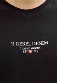 Μαύρο πουκάμισο με λευκό κείμενο «JJ REBEL DENIM CLASSIC GOODS EST.» και ένα μικρό κόκκινο τετράγωνο με το γράμμα «R» και το «2023» από κάτω.